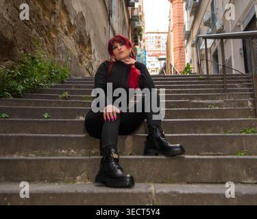 Giovane donna con capelli rossi tinti e tuta nera seduta sulle scale della città, trasuda fiducia nella moda urbana Foto Stock