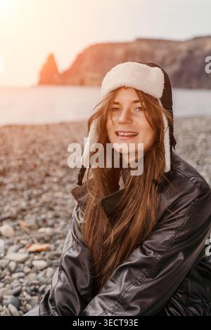 Donna spiaggia inverno giovane donna felice che indossa un caldo cappello ushanka seduto sulla spiaggia di ciottoli vicino al mare al tramonto Foto Stock