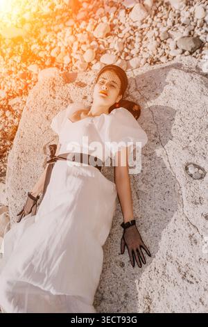 Donna abito bianco costa elegante giovane donna in abito di lino e guanti in pizzo appoggiati su pietre ruvide durante l'ora d'oro Foto Stock