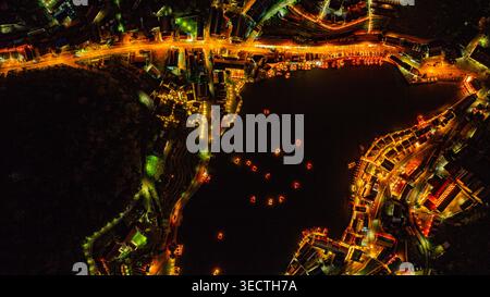 Una splendida vista aerea di un villaggio sul lago di notte, illuminato da calde luci dorate. Lanterne e barche galleggianti creano un modello ipnotico Foto Stock