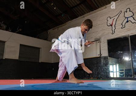 Uno studente di karate che indossa una cintura bianca e blu dimostra una posizione bassa e suggestiva sui tappetini da allenamento rossi e blu in un dojo. L'immagine acquisisce la messa a fuoco Foto Stock