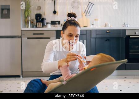 La mamma si siede sul pavimento e dà da mangiare a un bambino piccolo con un cucchiaio Foto Stock