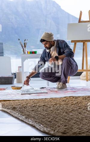 Artista maschile accovacciato sul pavimento in studio, dipingendo con coltello, berretto e cuffie Foto Stock
