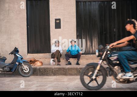 Mogotes, colombia - 21 marzo 2026 due uomini anziani che indossano cappelli seduti su gradini fuori da un edificio, osservano un giovane in moto sulla st Foto Stock