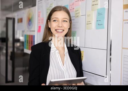 Donna in piedi accanto al tabellone di pianificazione in un ufficio moderno con un tablet Foto Stock