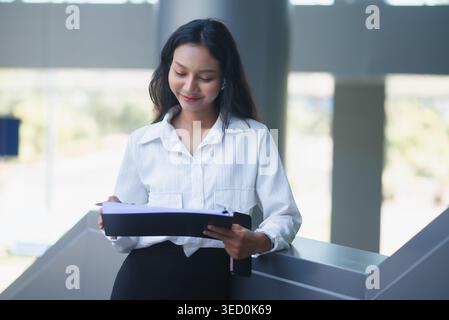 Una donna d'affari sorridente si trova accanto a una ringhiera, esamina documenti su un appunti e tiene in mano uno smartphone in un ufficio moderno, che rappresenta la produttività, Foto Stock