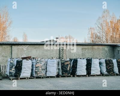 Grandi sacchetti sfusi pieni di legna da ardere tritata impilati su pallet in un cantiere industriale all'aperto. Ideale per i temi dell'energia da biomassa, della fornitura di combustibile, della logistica Foto Stock