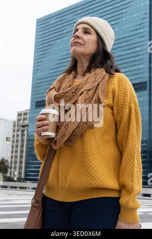 Donna adulta in piedi in piazza accanto a una torre di vetro blu, tenendo in mano una tazza di carta con coperchio bianco, guardando verso l'alto Foto Stock