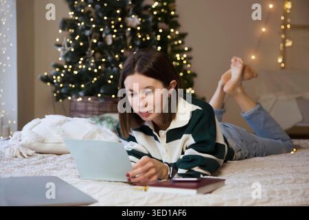 Donna scrive una lista di controllo di piani e sogni per il prossimo anno sdraiato sul letto di una casa nell'interno di Natale. Lista dei desideri per Natale. Pianificazione spese regalo. Obiettivi Foto Stock