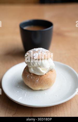 Semla - un tradizionale panino svedese di quaresima con cardamomo, panna montata e mandorle - su un piatto bianco con una tazza di caffè sullo sfondo Foto Stock