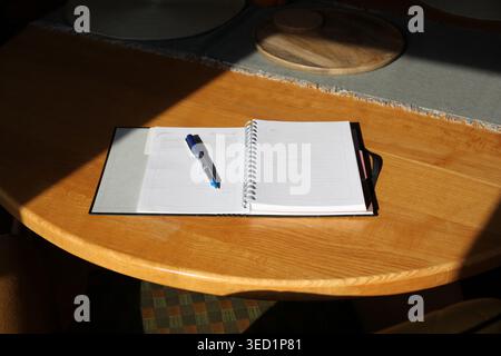 Pianificatore a spirale aperto con una penna blu su un tavolo rotondo di legno. Luce solare intensa che crea ombre forti in un ambiente accogliente. Foto Stock