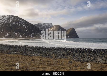 Le onde si infrangono sulle rive rocciose della spiaggia di Unstad, mentre le montagne innevate si innalzano drammaticamente contro il cielo nuvoloso, mostrando la valle incontaminata della natura Foto Stock