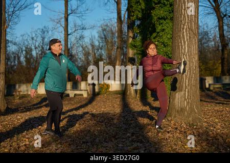 Donna di mezz'età che si appoggia in alto contro un tronco d'albero in un tratto, mentre il suo partner maschile guarda e sorride, riscaldandosi insieme prima di una corsa Foto Stock