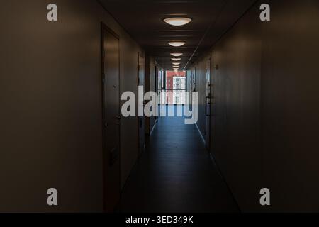 Il lungo e poco illuminato corridoio di un moderno edificio residenziale e' fiancheggiato da porte e illuminato da luci a soffitto che conducono ad una luminosa finestra. Questo Foto Stock