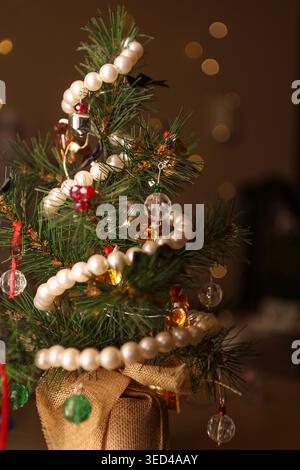 Un piccolo albero di Natale artificiale sorge in un caldo ambiente interno, adornato da perline e luci colorate, creando un'atmosfera festosa per il santo Foto Stock