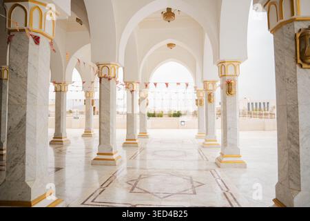 Colonne e archi in marmo bianco con dettagli dorati in un cortile aperto con pavimento geometrico e bandiere appese. Religione islamica, arco Foto Stock