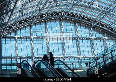 Interni della stazione internazionale di St Pancras con l'iconico tetto ad arco e scale mobili passeggeri nell'atrio principale. Due persone dalla silhouette sagomata salgono sulla scala mobile. Foto Stock