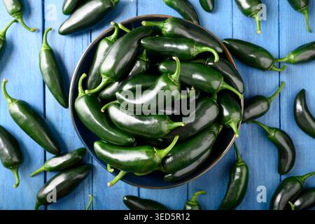 Recipiente con vista dall'alto pieno di peperoni jalapeno con peperoncino a caso su un tavolo di legno blu. Verdure fresche biologiche con pelle lucida e tonalità verdi vivaci Foto Stock
