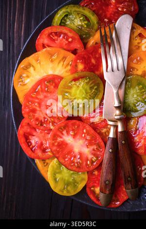 Pomodori cimellati disposti su un piatto scuro con forchetta e coltello. Ingredienti freschi biologici per insalate, colorato cibo vegetariano Foto Stock