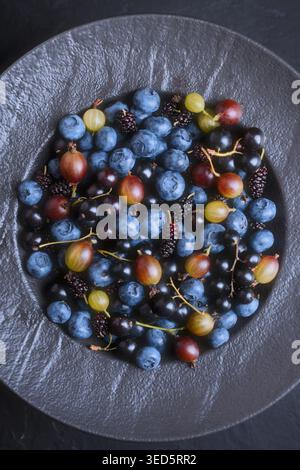 Mirtilli, spina e gelsi in una ciotola di ceramica scura su sfondo nero, vista dall'alto. Bacche blu fresche, stile alimentare minimale, sano concetto antiossidante Foto Stock