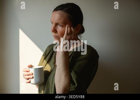 Profilo laterale di una donna di mezza età premurosa che tiene la tazza e tocca il viso guardando fuori dalla finestra, calda luce mattutina e atmosfera tranquilla e riflettente Foto Stock