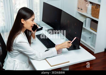 Giovane bella donna d'affari asiatica che chiama il broker, indicando con la penna un aumento del mercato azionario su pc per investire alti profitti in Modern Foto Stock