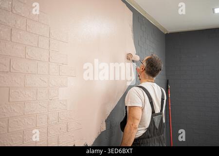 Un uomo sta usando un rullo per dipingere un muro di mattoni in questa scena. The mans Back è per noi, mentre applica un nuovo strato di vernice alla superficie grezza dei mattoni. Foto Stock