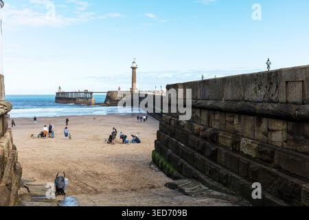Vista della spiaggia di Whitby nel North Yorkshire con gente che cammina e si rilassa sulla sabbia vicino alle mura del porto in pietra. Il molo e il faro si estendono Foto Stock