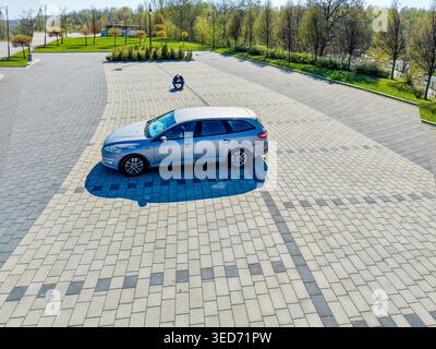 Cracovia, Polonia. Una station wagon grigia e argento parcheggiata in un ampio parcheggio e un anziano uomo dalla testa bianca seduto su un piccolo sgabello da pesca incentrato su una dro Foto Stock