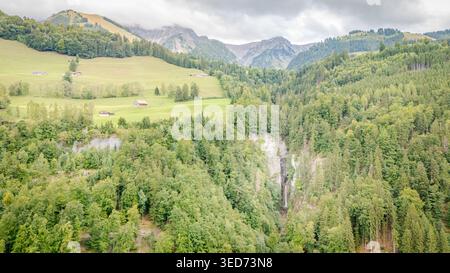 Vista aerea della cascata Grandvillard che si snoda attraverso una fitta foresta verde con prati alpini e vette di montagna sotto un cielo nuvoloso a Grandvillard Foto Stock