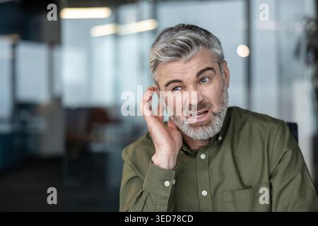 Uomo dai capelli grigi in un ufficio moderno che tocca l'orecchio e fatica a sentire, rappresentando concetti di perdita dell'udito, problemi di comunicazione e invecchiamento Foto Stock