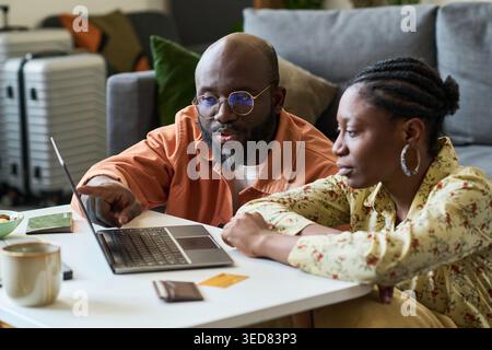 Coppia afroamericana che pianifica il viaggio con il computer portatile, controlla i dettagli della prenotazione e i documenti di viaggio a casa. Utile per la pianificazione delle vacanze, prenotazioni online, marketing del budget di viaggio Foto Stock