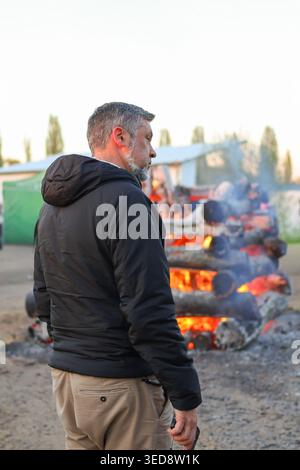 Un uomo felice di mezza età posa accendendo un falò in legno durante un evento all'aperto, catturando un'atmosfera accogliente e un momento di vita stagionale. Foto Stock