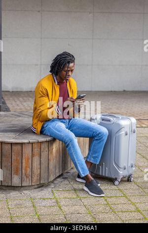 Uomo afroamericano seduto su una panchina di legno al plaza, giacca di senape, che controlla il telefono con la valigia Foto Stock