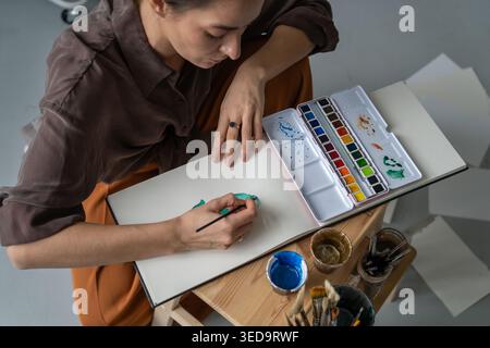 Artista donna rilassata che dipinge con acquerelli in un libro di schizzi a casa, tecnica di apprendimento e media. Foto Stock