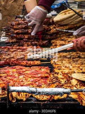 Una vivace scena di Street food ravvicinata con file di pollo e spiedini di carne conditi che grigliano su una grande superficie piatta. Un fornitore che indossa guanti utilizza tonnellate Foto Stock
