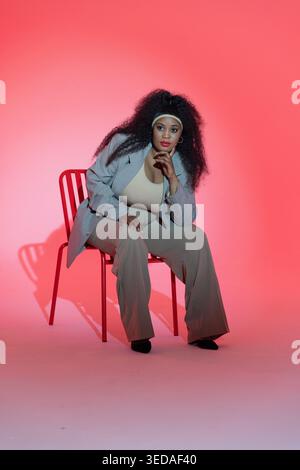 Donna seduta che indossa un blazer grigio chiaro che posa in studio con sedia e fascia in metallo rosso Foto Stock