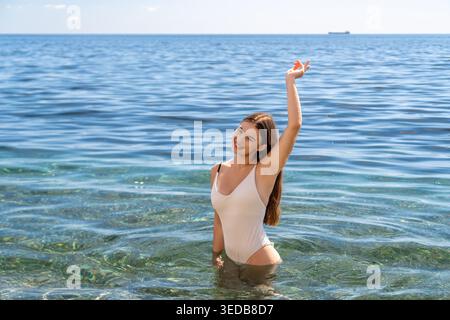 Costume da bagno donna mare sorridente giovane donna in costume bianco in piedi nel mare cristallino godendosi una vacanza estiva gioiosa Foto Stock