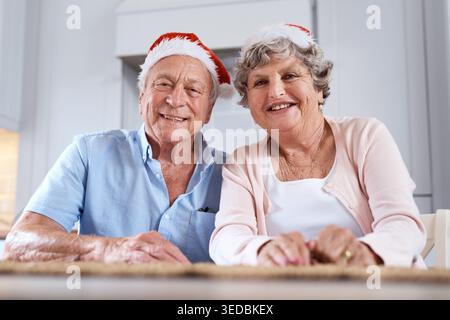 Natale, ritratti e coppie anziane in casa, sorridere e legare insieme alla tradizione festiva. Cappelli di Natale, uomo anziano e donna felice in cucina, amore Foto Stock