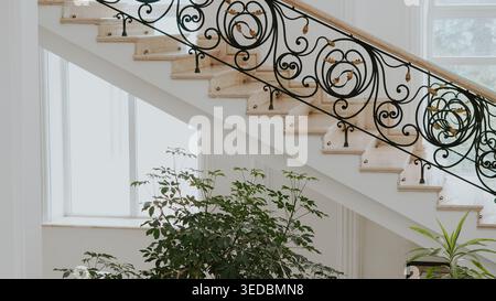 Elegante scalinata in marmo bianco con ringhiere ornate in ferro battuto in una sala illuminata dal sole, caratterizzata da lussureggianti piante interne verdi Foto Stock