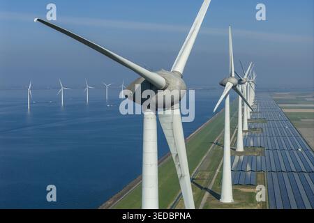 Vista aerea di un parco di grandi dimensioni con energia rinnovabile che presenta file di turbine eoliche e pannelli solari lungo una diga costiera sotto un cielo azzurro. Foto Stock