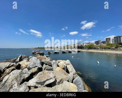 Panorama costiero con grandi rocce in primo piano e mare calmo che conduce a un lontano skyline urbano. Foto Stock