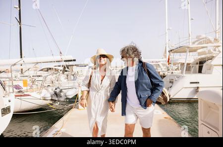 Felice coppia di anziani che si tiene per mano, godendosi una vacanza estiva passeggiando lungo il porto Foto Stock