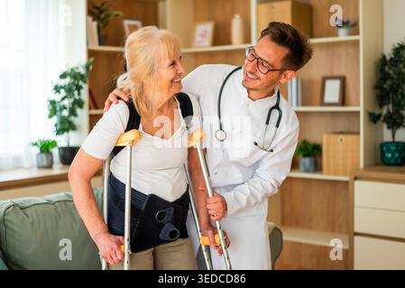 Un medico supporta una donna anziana che usa le stampelle, aiutandola a camminare in sicurezza a casa durante il recupero da una ferita. Foto Stock