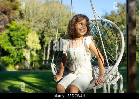 Preadolescente afroamericana seduta su una sedia in macrame in giardino tenendo la corda in una crema romper Foto Stock