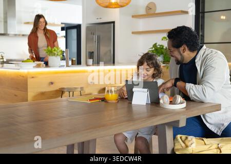 Diverse famiglie sedute al tavolo della cucina usando un tablet mentre la madre si trova sull'isola, copiando spazio Foto Stock