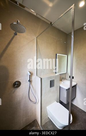 Bagno moderno con cabina doccia, WC a parete e lavandino compatto. Le piastrelle beige, il pavimento scuro e lo specchio illuminato creano interni essenziali e minimalisti. Foto Stock