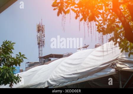 Una torre di telecomunicazioni sorge in modo prominente sopra il tetto di un edificio, dotata di antenne e parabole satellitari, contro un cielo azzurro. Foto Stock