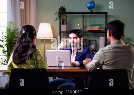 Coppia indiana in visita a un funzionario bancario con un laptop seduto sul tavolo durante la consultazione finanziaria discutendo i piani di investimento e immobiliari in modalità Foto Stock