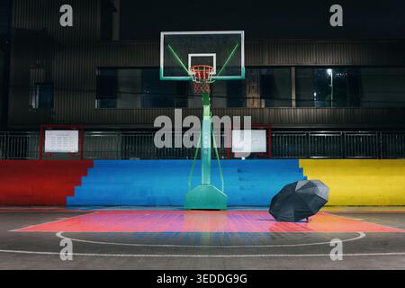 Campo da pallacanestro vuoto di notte con ombrello solitario appoggiato su un pavimento colorato e sport urbani Foto Stock